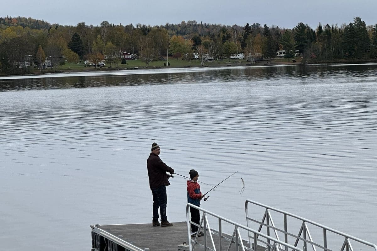 Two people fishing off the end of a dock in the fall, with autumn color along the far shore of the lake.