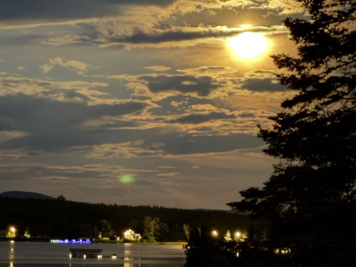 The sun low behind clouds over the lake, with lights coming on along the far shore.
