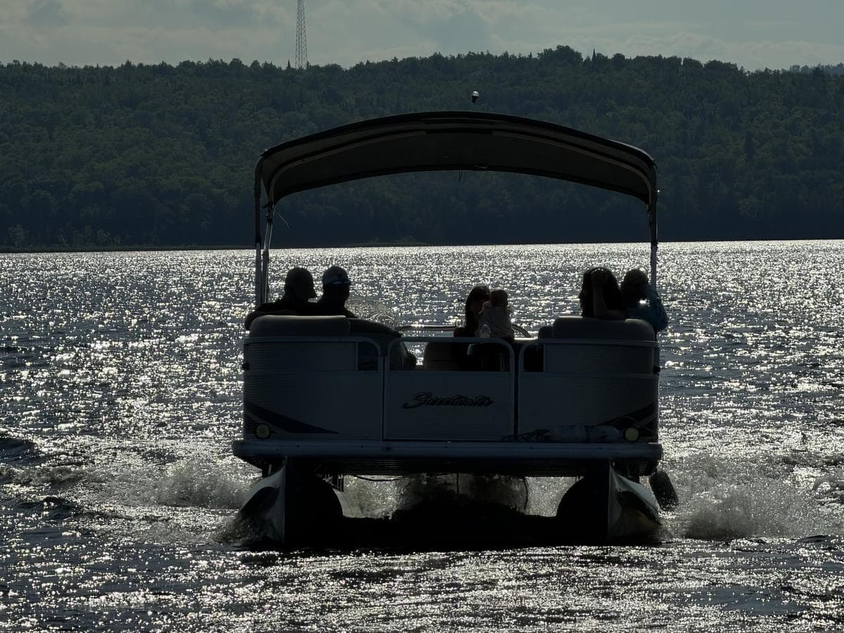A pontoon boat full of people heading out across sparkling water toward the wooded far shore.