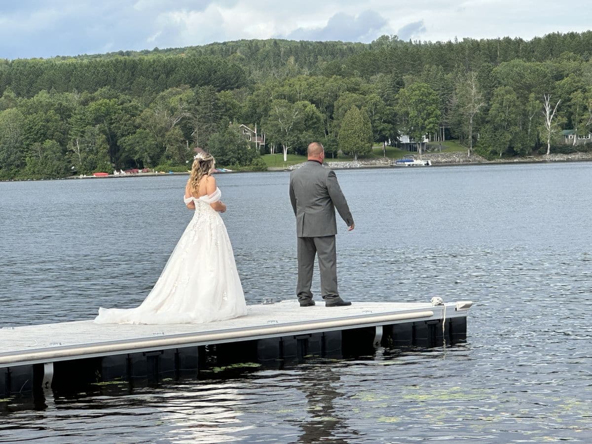 A bride and groom standing at the end of a dock, looking out across the lake to the wooded far shore.